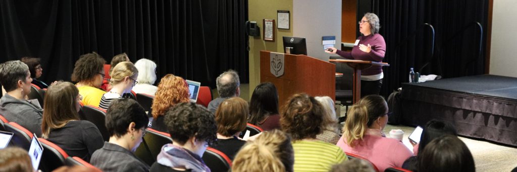 Abigail De Kosnik speaks from a podium in a lecture theatre. The audience is seen from behind at a three-quarter angle.