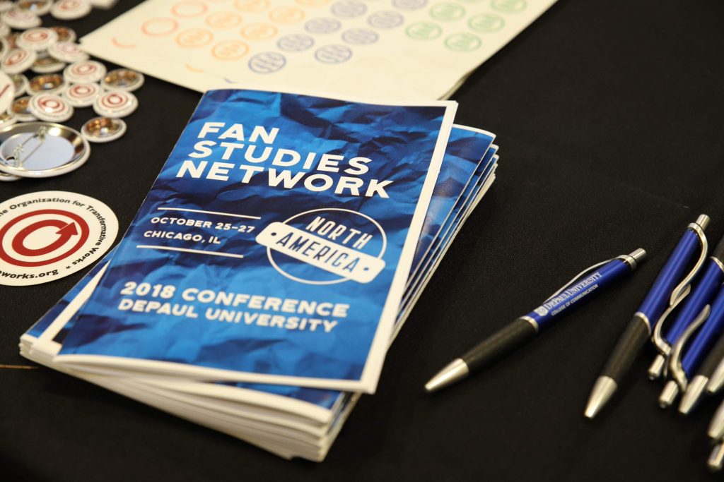 A stack of programs for the 2018 conference on a table next to some DePaul University–branded pens, stickers and buttons with the Organization for Transformative Works logo, and a sheet of pronoun stickers for people's badges.