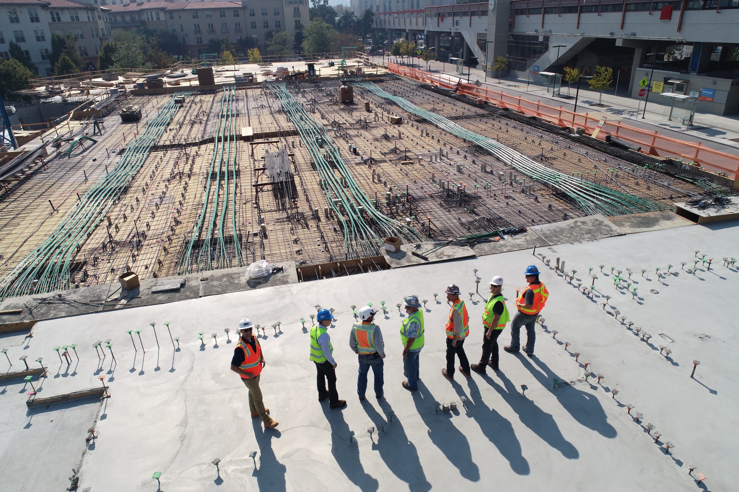 A line of workers in hard hats and high-vis vests standing on a concrete pad overlooking a large construction site. They cast long shadows behind them.